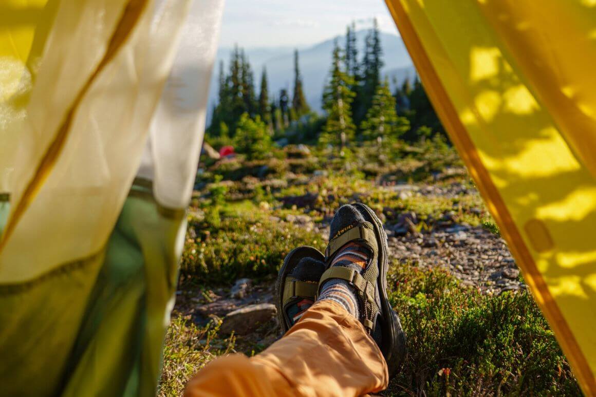 view of a person's feet wearing sandals poking out of a tent, with a view of the mountains and nature around them