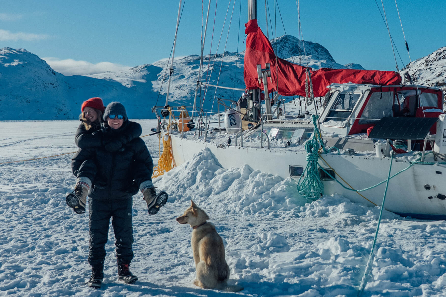 man, woman and dog in front of a sailboat on the ice