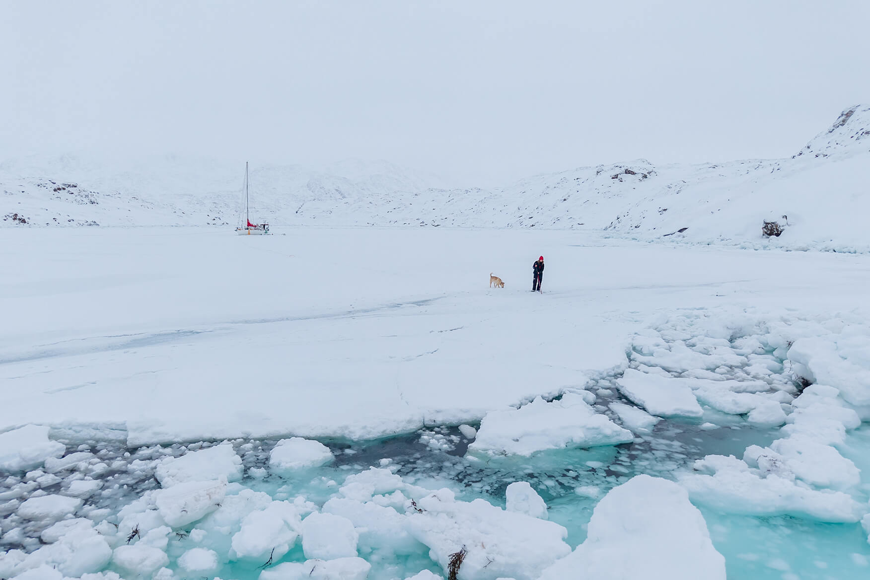 person and dog walking on ice with a sailboat in the background