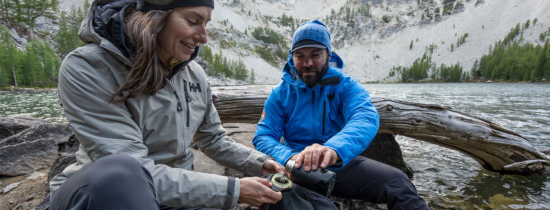 Two mountain guides stop for a break on their hike. The man pours water into the woman's bottle in the rugged mountain terrain.