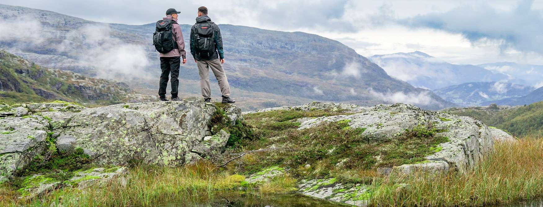 Woman hiking in Roam Cap, W Verglas 3L Shell Jacket, W Verglas 3L Shell Pant; man in Verglas 2L Shell Jacket, Tjern Tur Pant, Resistor Backpack.