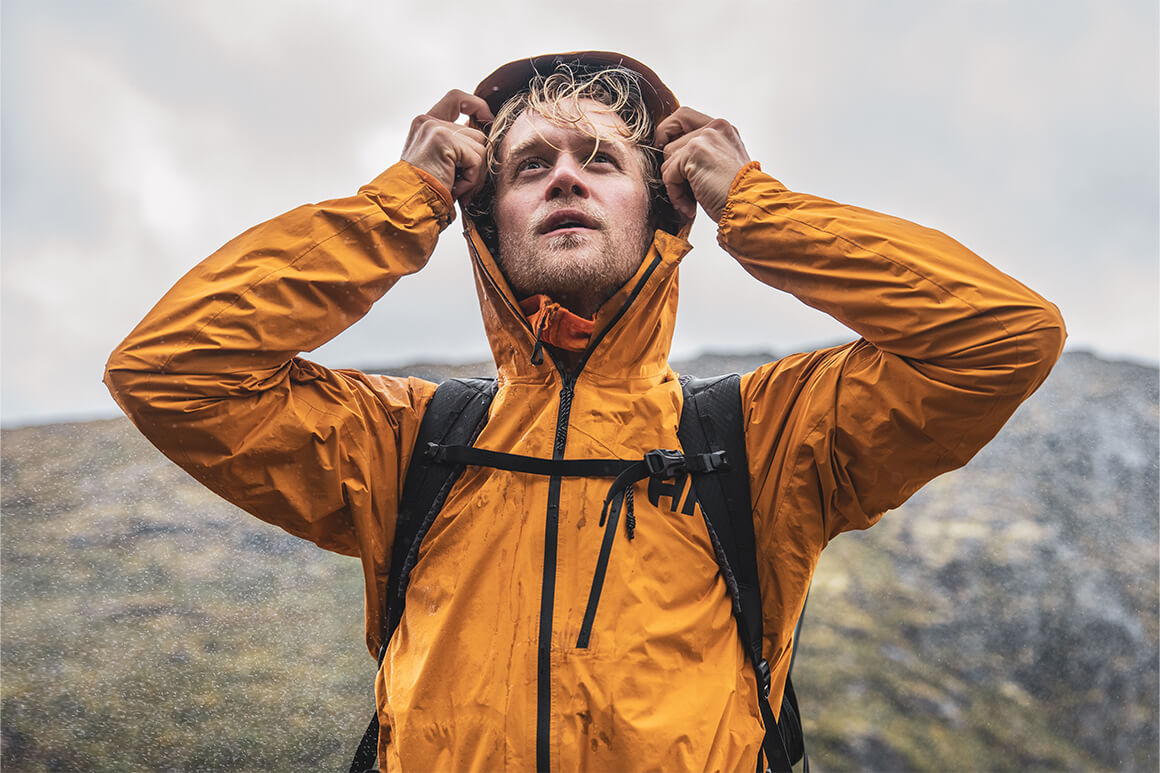 Man in an orange shell jacket putting on his hood in the mountains